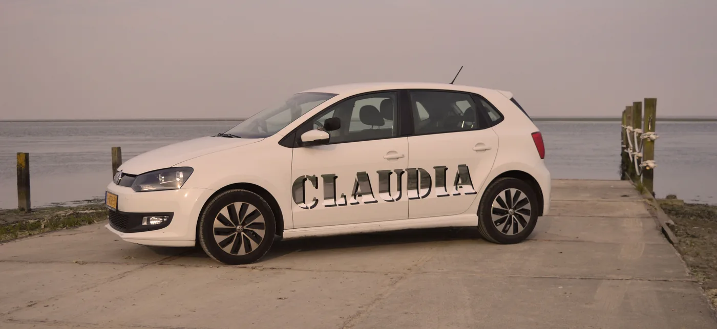 De witte lesauto met Claudia op de zijkant, geparkeerd aan het water